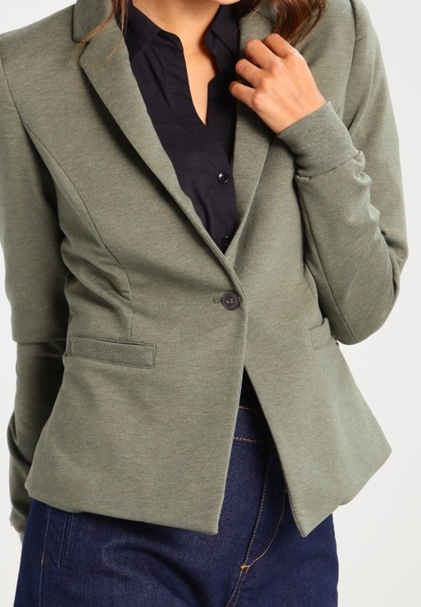 Femme portant un blazer vert olive ajusté par-dessus une chemise noire boutonnée et un jean bleu foncé, la main près du col.