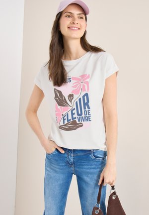 Ung kvinde iført hvid t-shirt med grafik af pink blomster og teksten "Fleur de Vivre", blå jeans, pink kasket, smilende, holder en brun taske.