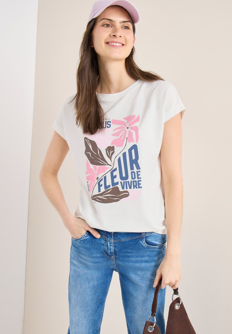 Jeune femme portant un t-shirt blanc à motifs avec des fleurs roses et le texte "Fleur de Vivre", jean bleu, casquette rose, souriante, tenant la sangle d'un sac marron.