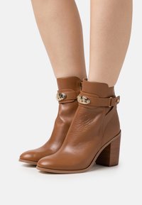 Botas de cuero marrón hasta el tobillo con punta redonda, tacón bajo y acentos de metal dorado, con una cremallera lateral y un tacón texturizado.