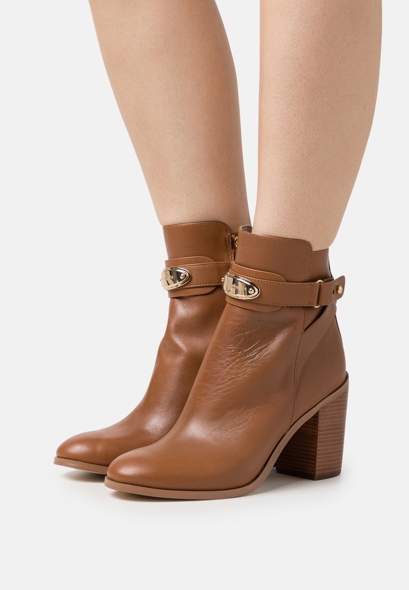 Botas de cuero marrón hasta el tobillo con punta redonda, tacón bajo y acentos de metal dorado, con una cremallera lateral y un tacón texturizado.