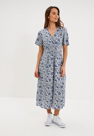 Femme portant une robe midi fleurie bleue et blanche à manches courtes et des baskets hautes blanches, debout devant un fond uni.