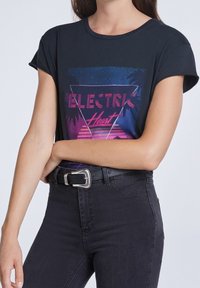 Camiseta negra de manga corta con mangas enrolladas que presenta un diseño gráfico colorido que incluye la palabra "ELECTRIC" y siluetas de palmeras, combinada con unos pantalones vaqueros negros de cintura alta y un cinturón con hebilla plateada.