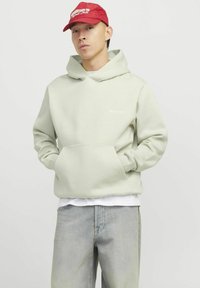 Hellgrüner Hoodie mit einer Fronttasche und Kapuze mit Kordelzug. Bestickter Text auf der Brust. Kombiniert mit hellblauen Jeans. Rote Mütze getragen.