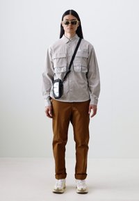 Light gray button-up shirt with multiple pockets, brown pants, and white sneakers. Complemented by black crossbody bag and sunglasses.