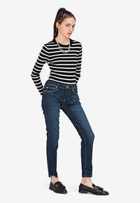 Czarna i biała w paski bluzka z długim rękawem połączona z ciemnoniebieskimi rurkami. Modelka nosi czarne loafersy z akcentem w postaci kokardy oraz naszyjnik z pereł.