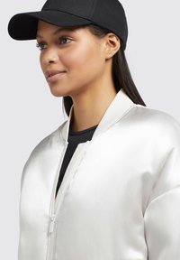 Giacca bomber in raso bianco con colletto a costine, chiusura con zip e cappellino nero; evidenziata la texture liscia e la vestibilità su misura.
