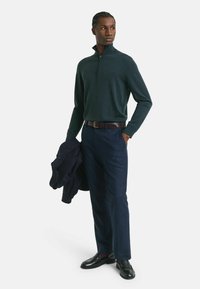 Dunkelgrüner Pullover mit Reißverschluss, marineblaue Hose, brauer Gürtel, schwarze Slipper, hält eine dunkle Jacke. Glatter Stoff, taillierte Passform, modernes Aussehen.