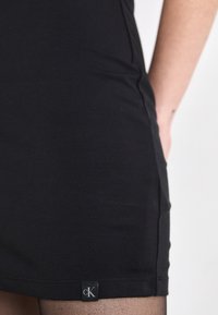 Black fitted dress made of smooth fabric, featuring a small CK logo tag at the hem. Simple design with no visible patterns or accents.
