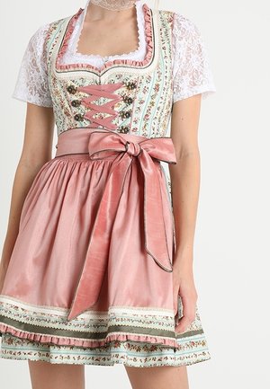 Kvinde iført en traditionel kjole med blomsterprint, blonder på ærmerne, lyserødt satinforklæde bundet i en sløjfe ved taljen og dekorative blondeaccenter.