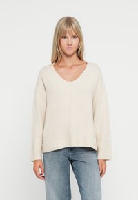 Beige übergroßer Pullover mit V-Ausschnitt, gerippten Bündchen und Saum. Weiche Textur mit leicht verkürztem Design. Kombiniert mit hellblauen Jeans.