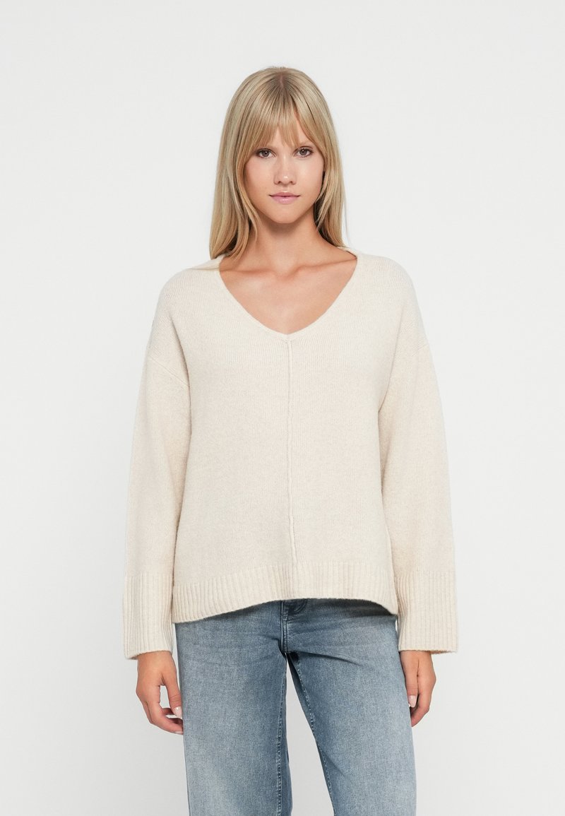 Beige übergroßer Pullover mit V-Ausschnitt, gerippten Bündchen und Saum. Weiche Textur mit leicht verkürztem Design. Kombiniert mit hellblauen Jeans.