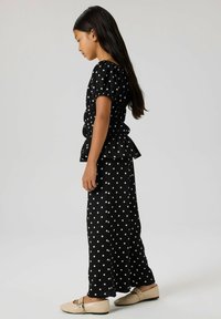 Crna majica s bijelim točkicama, balon rukavima i peplum rubom, uparena s odgovarajućim dugim hlačama. Mekani materijal, opušteni kroj, nošeno s cipelama svijetle boje.