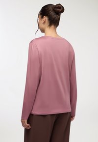 Koszula z długim rękawem w stonowanym różowym kolorze, z okrągłym dekoltem, gładką teksturą i luźnym krojem. Noszona z brązowymi spodniami.
