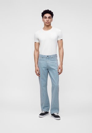 Jeune homme debout portant un t-shirt blanc uni, un jean bleu clair et des baskets noires sur un fond clair uni.