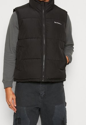 Homme portant un gilet matelassé noir avec fermeture éclair et poches latérales, par-dessus une chemise à manches longues grise et un pantalon cargo foncé.