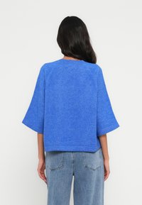 Pull bleu texturé à manches courtes avec une coupe décontractée, présentant des manches raglan et un col rond, associé à un jean en denim clair.