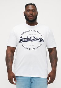 Weißes Baumwoll-T-Shirt mit einem auffälligen schwarzen Grafiklogo, das "Jack & Jones Denim Supply Co." auf der Brust zeigt. Kurze Ärmel, Rundhalsausschnitt.