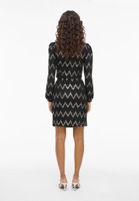 Rochie neagră cu model zigzag alb și gri, mâneci lungi bufante, talie ajustată și lungime scurtă. Modelul poartă sandale albe cu toc.