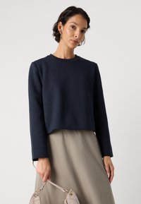 Sudadera corta azul marino con mangas largas y escote redondo, combinada con una falda beige. La modelo lleva un bolso gris claro.