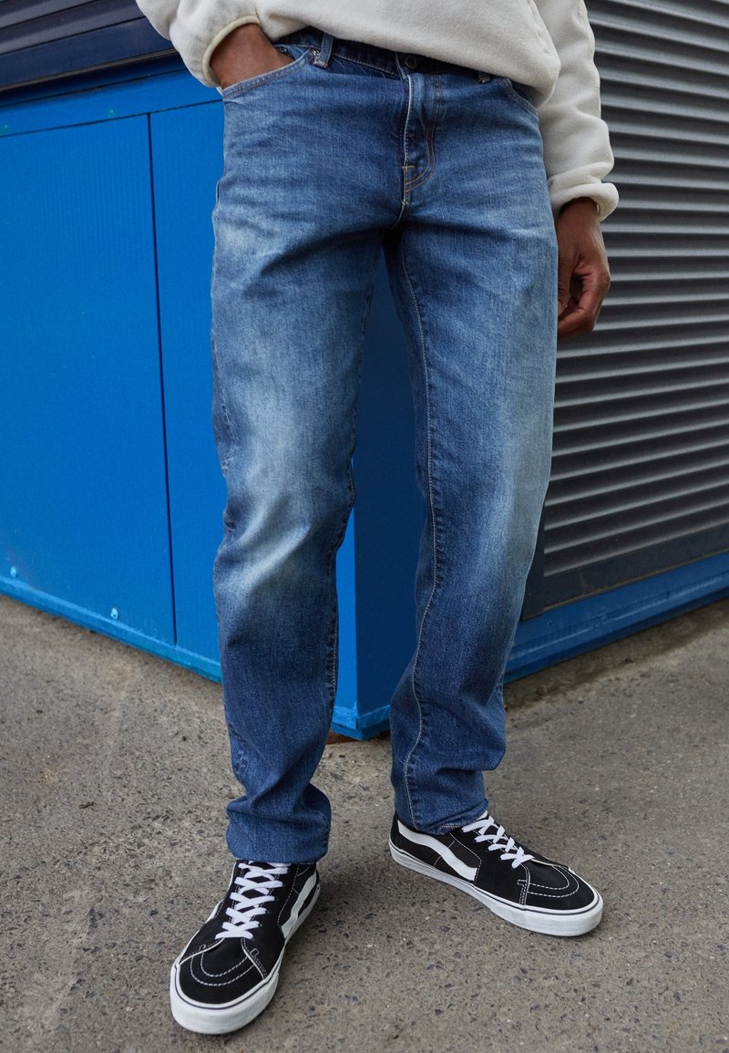 Vaqueros de mezclilla azul con lavado claro, ajuste recto, que presentan un diseño estándar de cinco bolsillos. Combinados con zapatillas negras con cordones blancos.