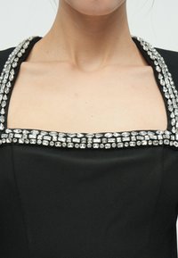 Μαύρο ένδυμα με τετράγωνη λαιμόκοψη διακοσμημένη με σειρά από καθαρά, λαμπερά στρας κατά μήκος των ραφών των ώμων, με ομαλή υφή.