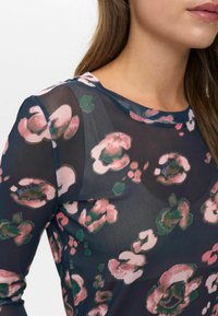 Donna che indossa un top trasparente blu notte con motivi floreali rosa e verdi, mostrando parzialmente il viso, il collo e la spalla.