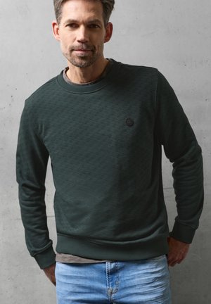 Mann mit kurzen Haaren und leichtem Bart, der einen dunkelgrünen, strukturierten Pullover und hellblaue Jeans trägt, steht vor einer grauen Wand.