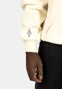 Felpa crema con polsini a coste, decorata con un logo grigio a forma di diamante sulla manica. Il tessuto sembra morbido e leggermente cangiante.