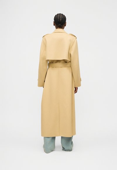 Άτομο με πλεγμένα μαλλιά, φορώντας μια μακριά μπεζ παλτό με ζώνη και ανοιχτή πλάτη, συνδυασμένη με ανοιχτό μπλε τζιν παντελόνι με φαρδιά πόδια, στεκόμενο με την πλάτη.