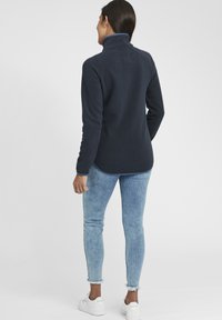 Pullover en polaire marine avec un col haut, présentant une texture douce et une bordure bleu contrastant. Associé à un jean ajusté bleu clair.