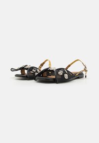 Musta slingback-sandaalit joustavalla pohjalla, jossa on metallisia koristeita ja teksturoituja hihnoja. Säädettävä nilkkahihna ruskeassa värissä.