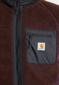Brūns flīsa žakets ar melnu neilona priekšpusi, ar rāvējslēdzēju un krūšu kabatu, kurai ir Carhartt logotipa uzlīme. Mīksta, strukturēta auduma.