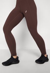 Leggings deportivas marrones de ajuste ceñido, con un diseño de costura sutil y un logo blanco. Combinados con zapatillas de correr negras.