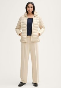 Doudoune beige au design matelassé, avec capuche et poches avant, portée sur un top marine et un pantalon large beige. Texture lisse, fermeture éclair.