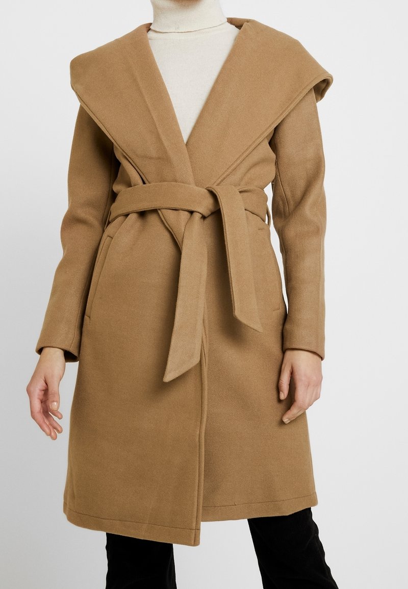 Femme portant un manteau en laine camel ceinturé sur un col roulé crème, debout les bras détendus le long du corps sur un fond uni.