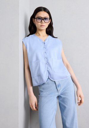 Jeune femme portant de grandes lunettes de soleil carrées, un haut sans manches bleu clair boutonné et un jean taille haute bleu clair, appuyée contre un mur.