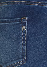 Tecido de denim em azul escuro com um acabamento texturizado, apresentando um pequeno bolso com um detalhe metálico e costura amarela em contraste.