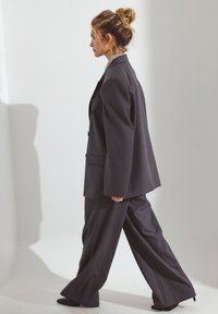 Tummanharmaa räätälinpuku, johon kuuluu väljempi bleiseri suurella kauluksella ja leveät housut. Sileä kangas, jossa on hienovarainen kiilto.