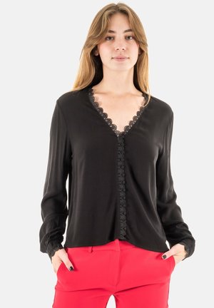 Blusa nera con scollatura a V, caratterizzata da dettagli in pizzo lungo i bordi. Maniche lunghe con polsini arricciati. Il modello posa con le mani nelle tasche.