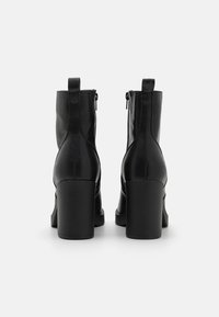 Paire de bottines en cuir noir avec talons carrés et languettes à l'arrière, vues de dos sur un fond blanc.