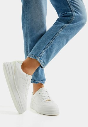 Personne portant un jean en denim bleu clair et des baskets blanches à plateforme, debout avec un pied légèrement levé.