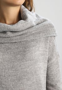 Jasnoszary, dziany sweter z szerokim, luźno układającym się dekoltem i fakturowaną tkaniną. Szczegół dekoltu prezentuje miękki, złożony wzór.