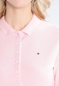 Jasnoróżowa koszulka polo z fakturowanej tkaniny, z kołnierzykiem, pięcioma guzikami i małym haftowanym logo na lewej piersi.