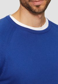 Blauer Rundhalsausschnitt-Pullover mit strukturiertem Rippmaterial. Hat einen weißen Innenkragen, der am Ausschnitt sichtbar ist, und kontrastierende Nähte.