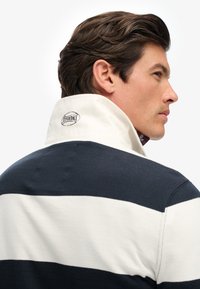 Marineblauwe en witte gestreepte polo met een hoge kraag. Bevat een verhoogd 'Superdry' logo op de kraag en is gemaakt van zachte katoen.