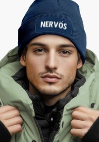 Navy knit beanie with "NERVÖS" patch, worn with an olive green puffer jacket. Textured fabric visible, with a black inner layer.