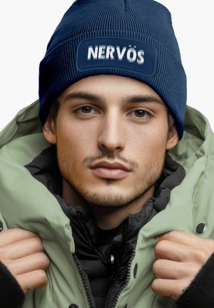 Navy knit beanie with "NERVÖS" patch, worn with an olive green puffer jacket. Textured fabric visible, with a black inner layer.