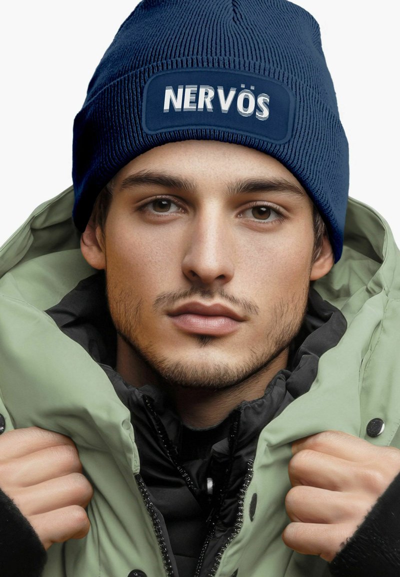 Navy knit beanie with "NERVÖS" patch, worn with an olive green puffer jacket. Textured fabric visible, with a black inner layer.