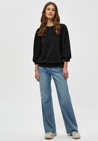 Črna trenirka z napihnjenimi rokavi, rebrastim robom; v kombinaciji z svetlobluzimi širokimi kavbojkami in bež teniskami. Nevtralna ozadje.